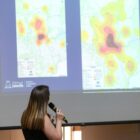 Mulher está virada para telão, de costas para a câmera enquanto faz apresentação