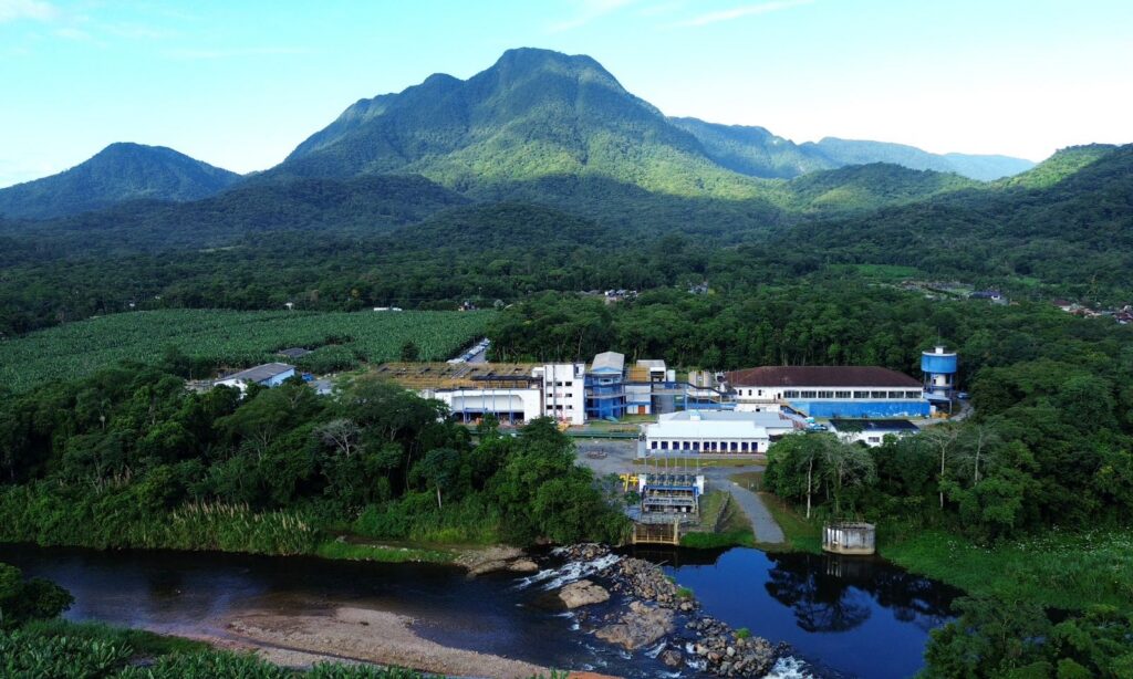 Imagem aérea mostra grande edificação da ETA Cubatão, nas cores branca e azul, envolta por um rio, montanhas e uma grande área de mata