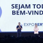 Homem e mulher brancos estão em pé, em um palco, com um painel com a frase sejam todos bem-vindos ao fundo