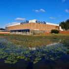 Imagem mostra prédio da Expoville ao fundo com lago com vegetação à frente