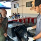 Homem alto, usando boné e luvas, mostra dois peixes vivos em bandeja para um casal de idosos negros