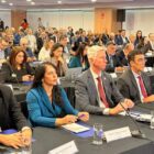 Imagem mostra homens e mulheres sentados lado a lado em mesas posicionadas uma atrás da outra em um grande auditório