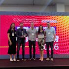 Cinco pessoas, duas mulheres e três homens, estão em um palco com um telão digital no fundo