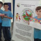 Três meninos de camisa azul estão em pé, perto de um banner sobre um projeto escolar. Um dos meninos segura um certificado, enquanto outros dois seguram um troféu