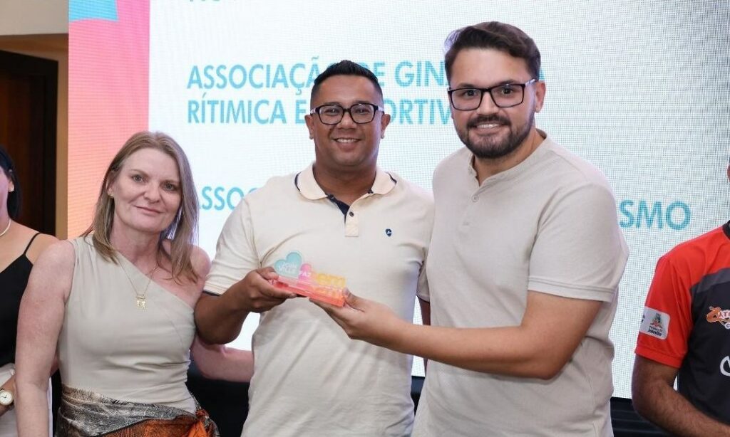 Uma mulher e dois homens estão em pé e sorriem para a foto. Os dois homens seguram o troféu do Joinville Faz Bem