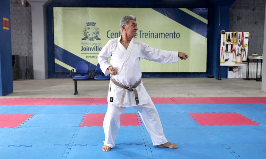 Homem idoso está vestido com kimono branco e faixa cinza e faz um movimento de karatê