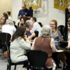 Homens e mulheres estão sentados distribuídos em duas mesas. Eles conversam e sorriem.