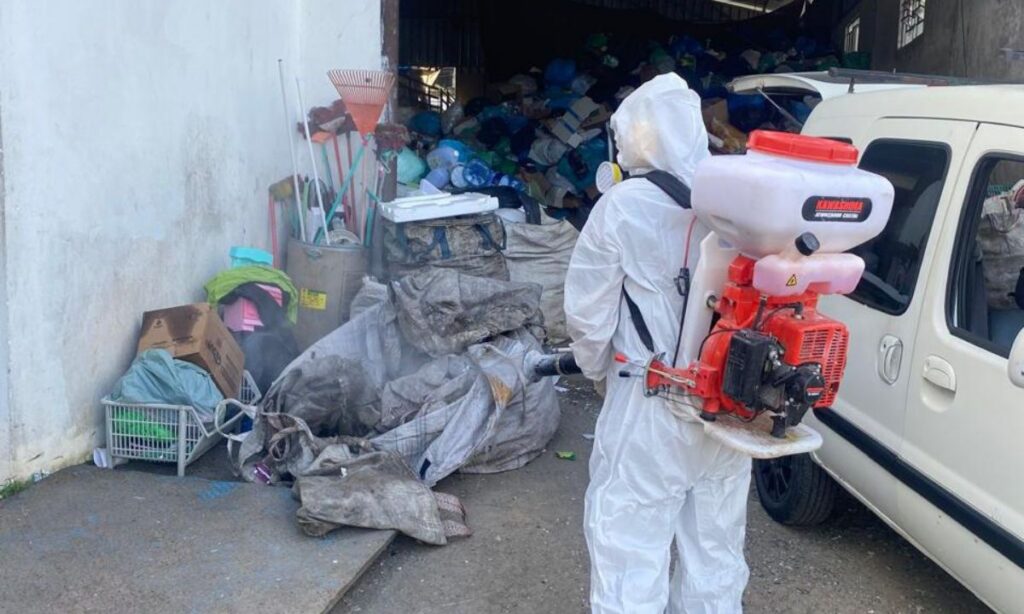 Pessoa vestida totalmente de branco, com equipamento de proteção, está usando equipamento de pulverização em área de entulho