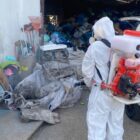 Pessoa vestida totalmente de branco, com equipamento de proteção, está usando equipamento de pulverização em área de entulho