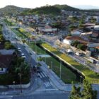 Imagem aérea de um passeio publico, com duas áreas de grama nas margens de um córrego, parquinho e outros equipamentos