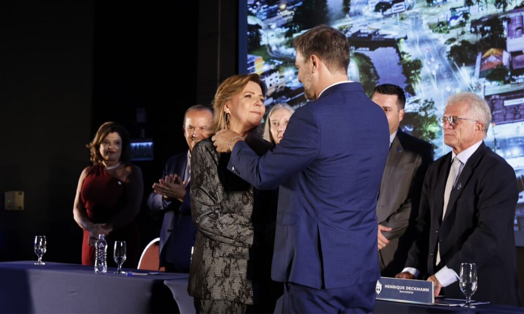 Um homem de terno virado de costas para a câmera coloca um botton na roupa de uma mulher loira que aparece emocionada