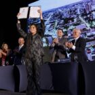 Mulher loira está em pé e ergue um documento para o alto com os dois braços enquanto sorri. No fundo, três homens e uma mulher aplaudem