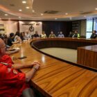 Imagem mostra uma sala ampla, com uma mesa oval ao centro, e diversas pessoas sentadas em cadeiras ao redor dela em uma reunião