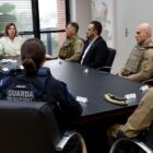 Imagem mostra pessoas sentadas em cadeiras ao redor de uma mesa. Uma mulher está na ponta e fala para os demais participantes da reunião, muitos deles com uniforme policial