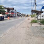 Imagem mostra rua pavimentada, sem calçada, com ponto de ônibus