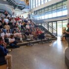 Homem em pé com microfone fala para plateia sentada a sua frente