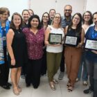 Quinze pessoas, entre homens e mulheres, posam para foto. Três delas seguram placas de reconhecimento
