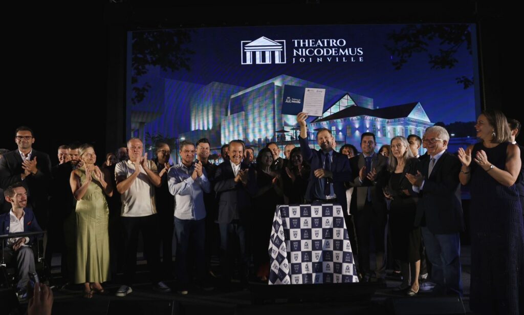 Um homem levanta um documento para o alto enquanto sorri. Ao seu lado, diversos homens e mulheres também sorriem e aplaudem