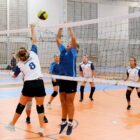 A imagem mostra mulheres jogando vôlei em uma quadra. Próximas à rede, duas delas se preparam para tocar a bola, que está no alto, enquanto as demais observam posicionadas