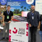 Quatro homens brancos posam para foto e sorriem em frente a um estande que com a descrição cidade de Joinville