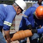 Homens uniformizados e com capacete manuseiam tubo usado na rede de esgoto