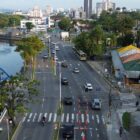 Imagem aérea mostra trecho de avenida pavimentada. Em um trecho há uma erosão no asfalto, que está sinalizada com cones