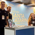 Quatro homens e uma mulher sorriem e posam para foto em um estande com painel sobre Santa Catarina