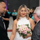 Homem de terno e mulher vestida de noiva, segurando buquê, estão a frente de um homem grisalho que usa óculos