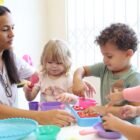 Mulher morena está acompanhando três alunos, uma menina e dois meninos, em uma mesa de um centro de educação infantil. As crianças estão brincando com brinquedos que remetem a uma cozinha, incluindo um mini fogão