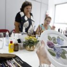 Em primeiro plano, a imagem mostra um prato de porcelana sendo pintado com desenho de flor. Ao fundo, há duas mulheres.