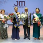 Na foto, há três homens e três mulheres lado e lado, em pé, posando para a foto e sorrindo. Eles seguram buquês de flores e estão com faixas comemorativas ao redor do pescoço.