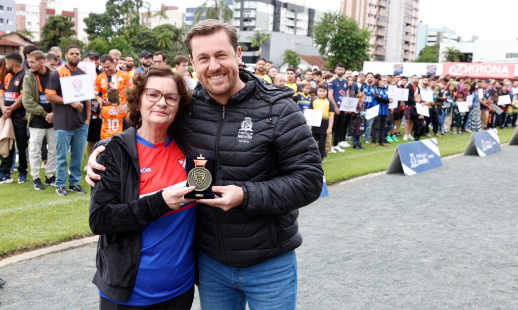 Um homem loiro e uma mulher idosa estão lado a lado, abraçados, e seguram juntos uma medalha