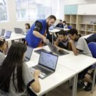 Em uma sala de aula, alunos estão sentados em cadeiras, em carteiras com notebooks. Um professor está em pé e orienta um dos alunos