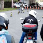 Em primeiro plano, há motociclistas de costas para a câmera. Ao fundo, há um motociclista fazendo manobras utilizando cones