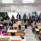 Imagem mostra sala de aula com crianças sentadas em carteiras e em pé. À frente, há vários adultos, homens e mulheres, em pé