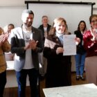 Imagem mostra homens e mulheres em pé, sorrindo e posando para fto. Uma mulher, ao centro, segura e exibe um documento