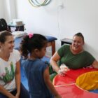 Em uma sala, uma mulher sorri para uma criança que está de costas, próxima a um brinquedo inflável. Outras duas mulheres na sala também sorriem.