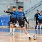 Imagem mostra quadra de futsal, onde menino com uniforme azul e preto se prepara para chutar uma bola. Ao fundo, há outros jogadores.