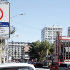 Rua com comércios e carros estacionados, destaque para uma placa de estacionamento rotativo