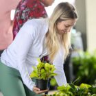 Mulher loira e jovem está abaixada e sorrindo enquanto segura um vaso de plantas na mão e escolhe outro