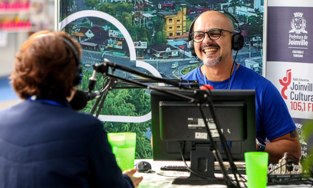 Imagem mostra homem com fone de ouvido sorrindo para pessoa que está à sua frente e fala em um microfone. Ambos estão sentados perto de uma mesa