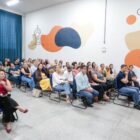 Imagem mostra homem de calça jeans e camiseta branca à frente de uma ampla sala, em pé, com um microfone na mão. À frente dele, há pessoas sentadas na plateia