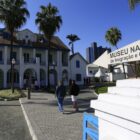 Imagem mostra o Museu Nacional de Imigração e Colonização, imóvel de dois andares em branco com detalhes em preto. Há uma seta indicativa com o nome do museu e algumas pessoas estão caminhando na calçada