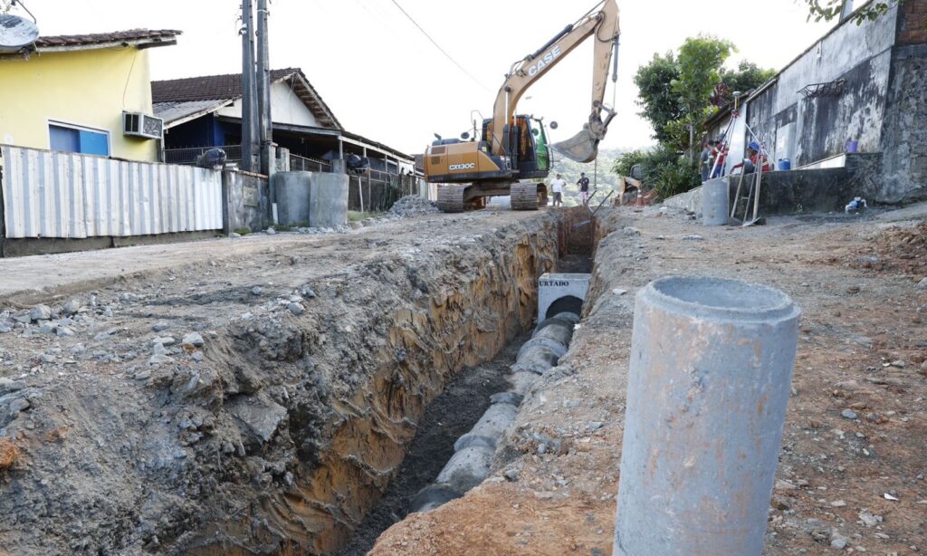 Máquina trabalha em obra de drenagem e pavimentação em rua de barro. Em uma vala, há tubos no solo