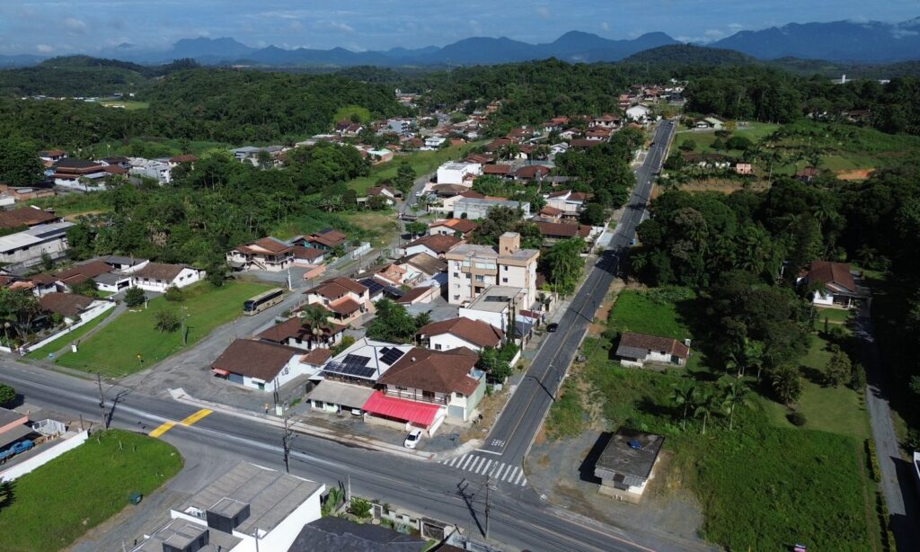 Imagem aérea de rua pavimentada em terreno elevado. No entorno, há moradias e vegetação.