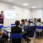 Em sala da aula, adolescentes uniformizados estão sentados em cadeiras ao redor de grandes mesas. à frente, um professor está em pé, falando com os alunos
