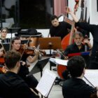 Imagem mostra ensaio de uma orquestra. Há músicos, homens e mulheres, vestidos de preto, tocando diferentes instrumentos. Uma mulher está em pé e sorri.