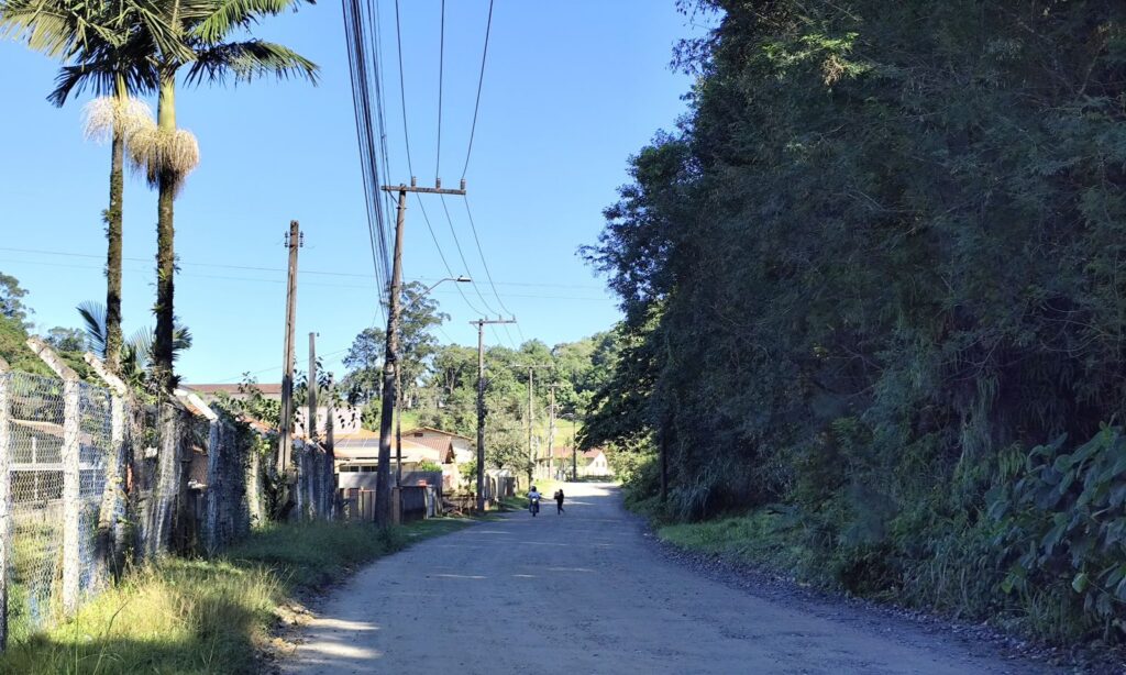 Em rua sem pavimentação, uma moto e uma mulher aparecem em movimento ao fundo. A rua é margeada por casas e vegetação.