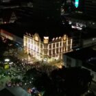 Imagem aérea noturna mostra grande prédio iluminado localizado em esquina no centro de Joinville