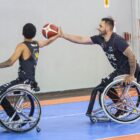 Dois homens estão sentados em cadeiras de todas em uma quadra de basquete. O paratleta posicionado mais à frente está recebendo a bola de basquete do outro jogador.